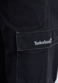 Close-up van zwarte stoffen broek met een Timberland-logo op de zak, waarbij de stiksels en textuur van het materiaal worden benadrukt.