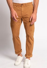 Khaki chinos o prostym kroju, z podwiniętymi mankietami, z zapięciem na guzik i przednimi kieszeniami, zestawione z białymi sneakersami na neutralnym tle.