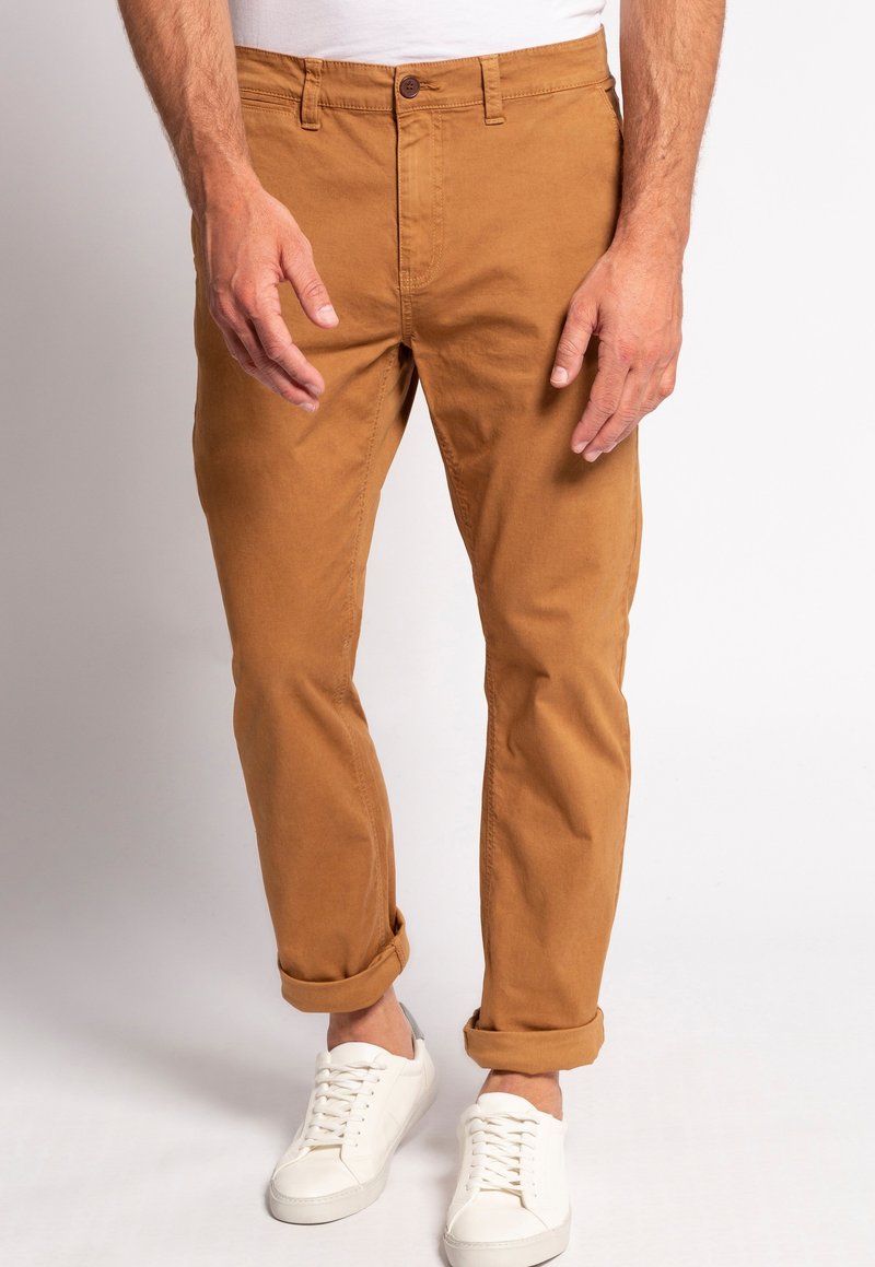 Khaki chinos med lige snit, opsmøgede bukseben, knaplukning og forlommer, parret med hvide sneakers på en neutral baggrund.