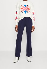 Gestreifter Pullover in Elfenbein mit mehrfarbigem geometrischen Muster und langen Ärmeln, kombiniert mit navyblauen Schlaghosen und weißen High-Heels Stiefeln.