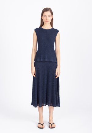 Žena stoji, nosi tamnoplavi teksturirani top bez rukava i odgovarajuću midi suknju s crnim sandalama otvorenih prstiju, na bijeloj pozadini.