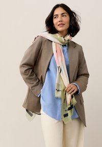 Blazer beige sopra una camicia azzurro chiaro, pantaloni crema, con una sciarpa di seta a motivi con tonalità rosa e verde, e bordi frangiati.