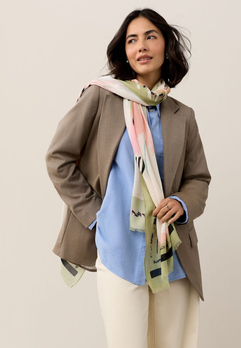 Blazer beige sopra una camicia azzurro chiaro, pantaloni crema, con una sciarpa di seta a motivi con tonalità rosa e verde, e bordi frangiati.