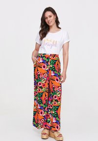 Weißes T-Shirt mit dem Text "think bright", kombiniert mit bunten, gemusterten Palazzo-Hosen mit blühenden Designs in Orange und Grün. Espadrille-Sandalen.
