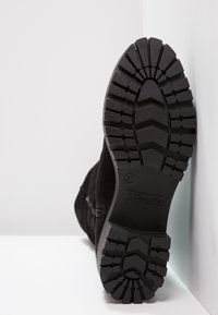 Botín negro de ante con una suela de goma texturizada que presenta un patrón de zigzag, cremallera lateral y logo de la marca en la suela.