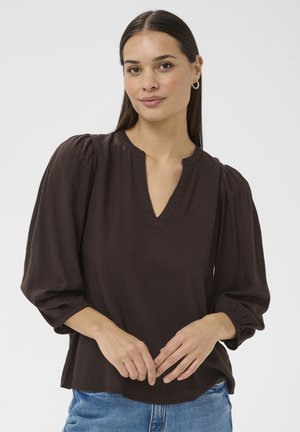 Kobieta o długich brązowych włosach, nosząca złote kolczyki koła, ciemnobrązową bluzkę z długim rękawem z dekoltem w serek oraz niebieskie dżinsy na białym tle.