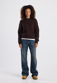 Brown ribbed sweater with a round neckline, worn over a white shirt, paired with loose blue jeans and tan shoes. Textured fabric detail visible.