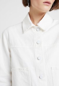 Biała kurtka dżinsowa z zapięciem na guziki, wystającym kołnierzem i dwiema przednimi kieszeniami. Tkanina ma teksturowane wykończenie z subtelnymi detalami w stichingu.