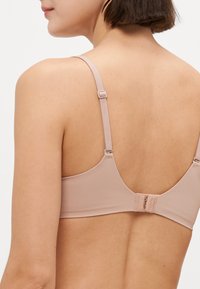 Woman wearing a light pink adjustable bra with thin straps and a hook-and-eye back closure, viewed from behind at shoulder height.