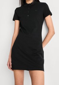 Femme portant une robe polo noire courte avec des manches courtes et un col boutonné, debout devant un fond clair uni.