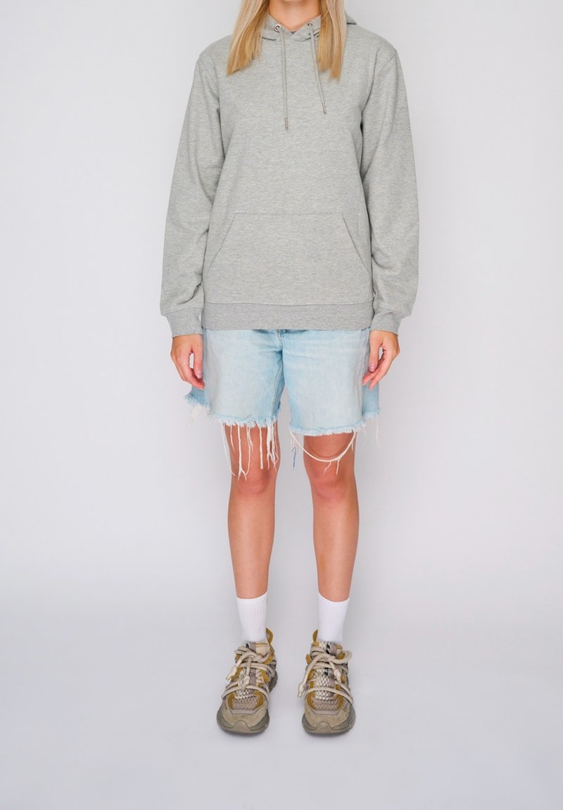 Personne portant un sweat à capuche gris, un short en jean bleu clair déchiré, des chaussettes blanches et des baskets beiges, debout devant un fond uni.