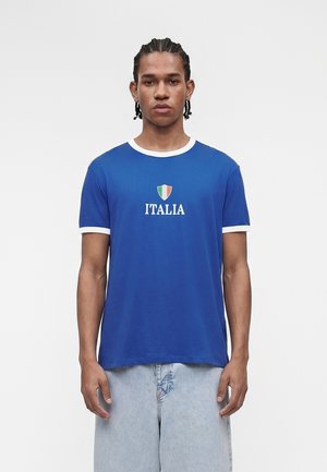 Νέος άνδρας με πλεξουδάκια, φορώντας μπλε μπλουζάκι με λευκή επένδυση και τη λέξη «ITALIA» κάτω από μια ασπίδα με την ιταλική σημαία, συνδυασμένο με ανοιχτόχρωμο τζιν.