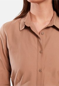 Camicia marrone con bottoni, dotata di colletto e due chiusure a bottoni, con una texture liscia e dettagli di cucitura minimi lungo il colletto.