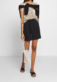 Blus med blomstertryck, kortärmad i gult och svart, kombinerad med svarta högmidjade shorts. Vit pärlhandväska; svarta sandaletter.