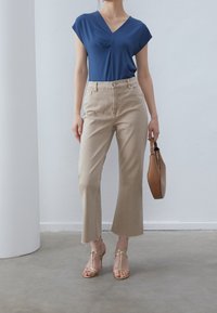 Kvinna iförd en blå V-ringad topp, beige vida cropped jeans, guldiga klackar med remmar, hållande en brun handväska i en minimalistisk inomhusmiljö.