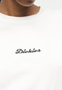 Κοντινό πλάνο ενός ατόμου που φοράει λευκό πουκάμισο με την επιγραφή «Dickies» κεντημένη με μαύρα γράμματα στο στήθος.