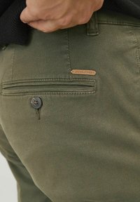Pantalones verde oliva con un acabado texturizado, que cuentan con un bolsillo trasero con un botón negro y una pequeña etiqueta de cuero marrón.
