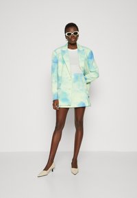 Blazer en tie-dye bleu pastel et vert assorti à une jupe. Présente une conception structurée et une texture superposée. Porté avec des lunettes de soleil blanches.