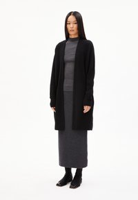 Schwarze Strickjacke über einem grauen gerippten Pullover, kombiniert mit einem dunkelgrauen Rock und schwarzen Stiefeletten. Glatte Texturen und minimalistisches Design.