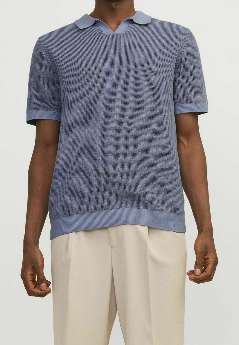 Polo tričko s krátkým rukávem, modré pletené s texturovaným vzorem, s žebrovaným límcem a manžetami. Nošeno s světlými, skládacími kalhotami.