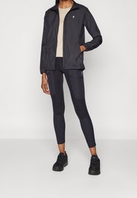 Veste légère noire à fermeture éclair, associée à des leggings noirs avec des détails en patch. Haut beige uni et chaussures de sport noires.