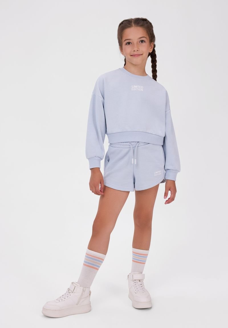 Jeune fille aux cheveux tressés, portant un sweat-shirt bleu clair et un short, des chaussettes rayées et des baskets blanches montantes, debout devant un fond uni.