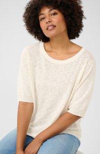 Un pull en tricot léger de couleur crème avec une coupe ample, un col rond large et des manches courtes. Accompagné d'un jean bleu, assis contre un fond gris.
