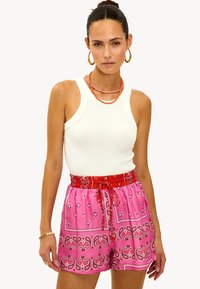 Top sin mangas blanco con pantalones cortos de patrón rosa que presentan una cinturilla elástica roja y cordón. Pendientes de aro dorados y collares de cuentas en capas.