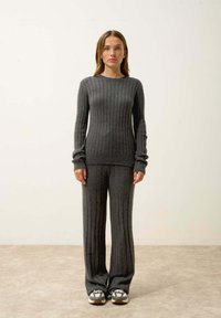 Ciemnoszary sweter z prążkowanej dzianiny z długimi rękawami i pasującymi prążkowanymi spodniami. Sneakersy w białe i szare akcenty, stojące na neutralnym tle.