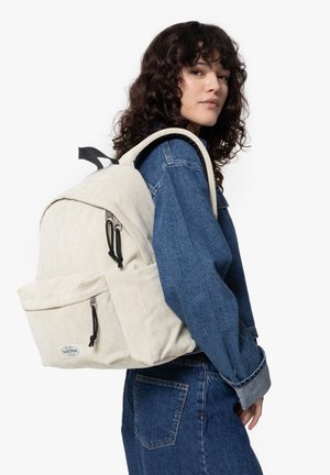 Sac à dos en velours côtelé beige clair avec des bretelles rembourrées, une poche avant et des tirettes de fermeture éclair noires. Porté sur le dos d'une personne en tenue en denim.
