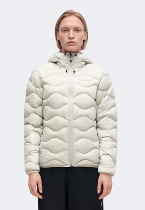 Femme aux cheveux blonds courts portant une veste matelassée à capuche gris clair et un pantalon noir, debout devant un fond uni.