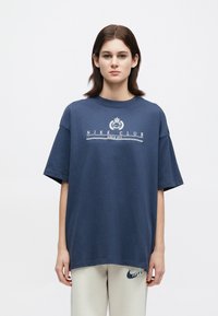 Νέος με μαλλιά έως στους ώμους σε σκούρο χρώμα, φοράει μπλε oversized μπλούζα Nike Club και εκρού παντελόνι με το λογότυπο της Nike.