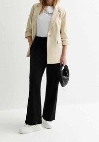Veste beige, t-shirt blanc avec texte noir, pantalon noir et baskets blanches ; tenant un sac noir tissé. Accent sur une coupe classique et décontractée.