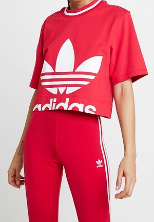 Osoba nosząca czerwoną krótką koszulkę Adidas z białym logo oraz dopasowane czerwone legginsy z białymi paskami i małym logo Adidas na udzie.