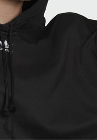 Sudadera negra de tela suave, con capucha ajustable y un logo de tres rayas blancas en el pecho. Superficie texturizada y corte amplio.