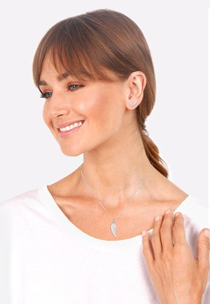 Collana d'argento con pendente a forma di ala e orecchini a chiodo abbinati. I gioielli sono indossati con una camicia bianca e presentano superfici lisce e lucide.