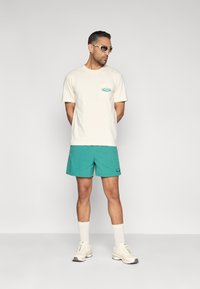 Ανοιχτό μπεζ μπλουζάκι με τσέπη στο στήθος, συνδυασμένο με τεϊλ σορτς. Αξεσουάρ περιλαμβάνουν ανοιχτόχρωμα γυαλιά και λευκές αθλητικές κάλτσες και παπούτσια.