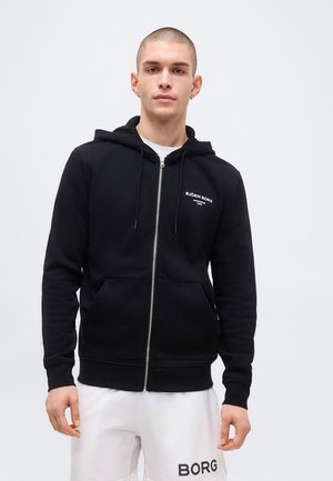 Jonge man die een zwarte hoodie met rits draagt met de tekst "BJÖRN BORG" en witte shorts met het "BORG" logo, staand tegen een effen lichte achtergrond.