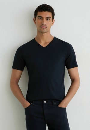 Jonge man met krullend haar die een zwart T-shirt met V-hals en korte mouwen en een donkere broek draagt, staand tegen een lichte muur met handen in de zakken.