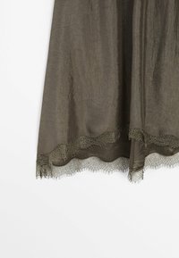 Gros plan d’un tissu vert olive avec une texture froissée et un galon en dentelle festonné le long du bord inférieur.