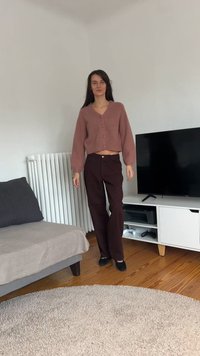 Ženska stoji v dnevni sobi, oblečena v kratki vijolični pulover in temne hlače, blizu sivega kavča in bele TV enote z igralnimi krmilniki.