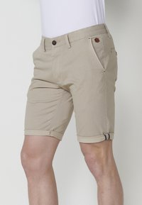 Beige shorts made from cotton blend fabric, featuring a rolled hem, two front pockets, and a brown button detail. Flat front design.