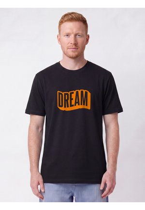 Homme aux cheveux roux courts portant un t-shirt noir avec le mot "DREAM" en lettres orange en gras, debout devant un fond uni.