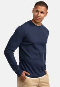 Marineblauwe ribgebreide trui met lange mouwen en een ronde halslijn, gemaakt van gestructureerde stof, gedragen over een lichtgekleurde blouse, gecombineerd met beige broek.