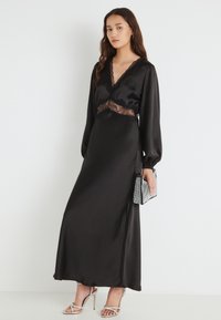 Čierne maxi šaty zo saténu s dlhými rukávmi, čipkovým detailom v páse, hlbokým výstrihom do V a bočným rozparkom, doplnené striebornou kabelkou.