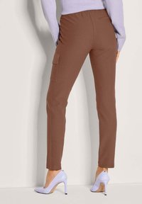 Frau in braunen, slim fit Cargo-Hosen mit Seitentasche und lila High-Heels, die an einer hellen Wand steht.
