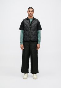 Chaleco acolchado negro sobre una camisa de manga larga verde, combinado con pantalones anchos acolchados negros y zapatillas beige. Diseño simple, ajuste moderno.