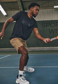 Juodas sportinis marškinėlis su "BOSS" logotipu, šviesiai rudos spalvos šortai su šoninėmis pūtimis ir pilki sportbačiai su juodais bei neoniniais akcentais, tenisisto pozoje.