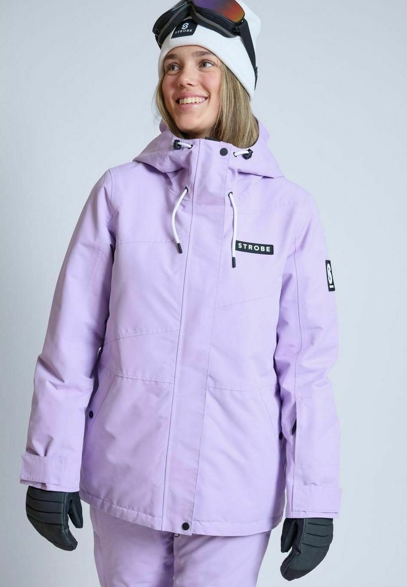 Lilla skijakke med justerbar hætte, frontal lynlås og logo-patch. Teksturstof og sidelommer, parret med sorte handsker og en hvid hue.