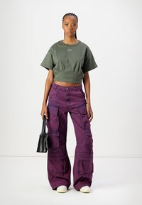 Persoon in paarse cargo broek en een groene t-shirt, die een kleine zwarte handtas vasthoudt tegen een plain witte achtergrond.